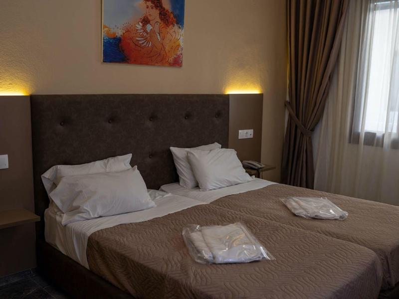 Zweibettzimmer mit braunem Kopfteil, beigen Bettdecken, weißen Kissen und zwei Fenstern mit Vorhängen.