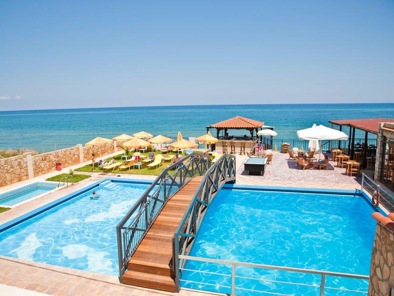 Blick auf zwei Pools mit einer Holzbücke, Sonnenschirmen und Meer im Hintergrund.