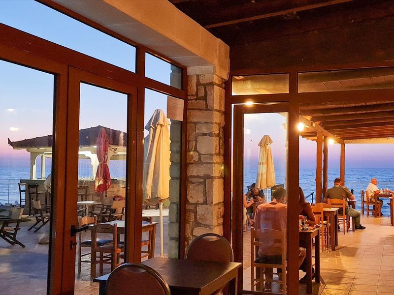 Restaurantterrasse mit Holzmöbeln, Steinmauern und Blick auf das Meer bei Sonnenuntergang.