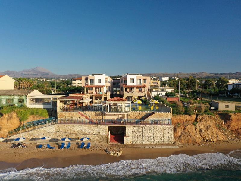 Strand mit blau-weißen Liegestühlen und Häusern auf felsigem Hügel bei klarem Himmel.
