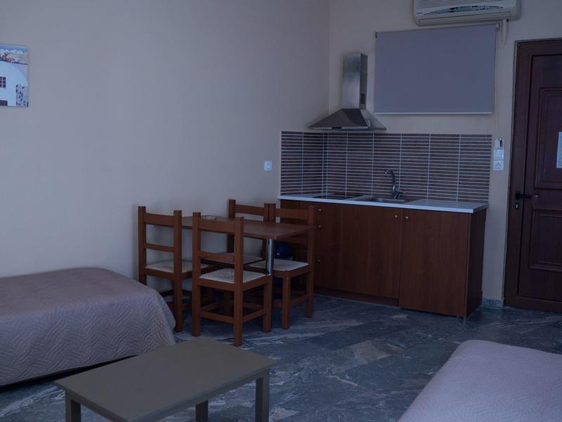 Kleines Zimmer mit zwei Betten, Holztisch, Stühlen, Küchenzeile und Tür. Wände hell, Boden Fliesen.