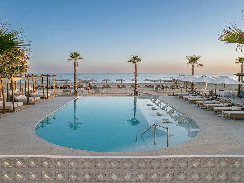 Zone de piscine avec chaises longues et palmiers, vue sur la plage et la mer sous un ciel clair.