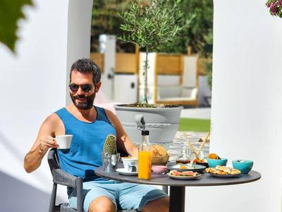 Hombre sentado al aire libre en mesa con desayuno bebiendo café frente a pared blanca con arco.
