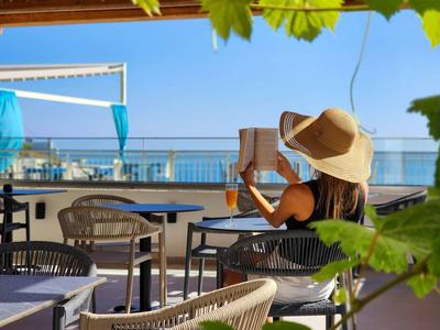 Persona con sombrero de sol leyendo un libro en terraza con vista al mar bajo el sol