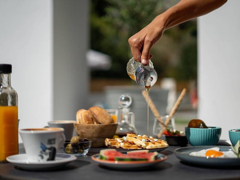 Hand giet vloeistof over eten op een tafel met ontbijtgerechten.