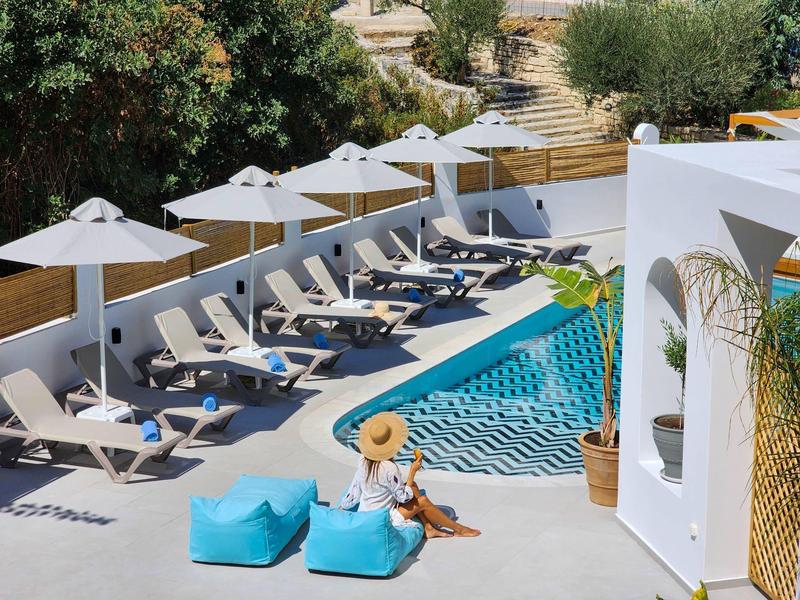 Vrouw met hoed zit naast blauw zwembad met ligbedden en parasols onder bomen.