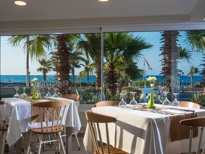 Restaurant met witte tafels en stoelen, uitzicht op palmbomen en zee door grote ramen.