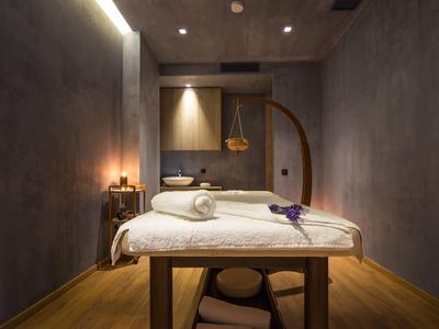 Schlichte Massageliege mit weißer Decke, Holzboden, graue Wände und gedämpftes Licht.