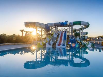 Wasserpark mit Rutschen in verschiedenen Farben neben großem, ruhigem Schwimmbecken bei Sonnenuntergang.