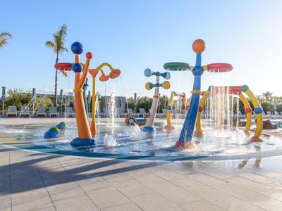 Bunter Wasserspielplatz mit Spritzdüsen, transparenten Wasserstrahlen und Palmen im Hintergrund.
