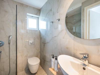 Helles modernes Badezimmer mit Marmorwänden, rundem Spiegel, Toilette und Waschbecken mit Armatur.