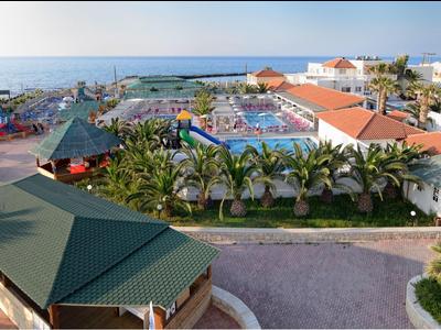 Ferienanlage am Meer mit Pool, Palmen, roten Dächern und umliegenden Gebäuden bei Sonnenschein.