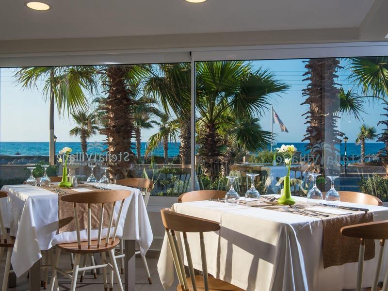 Restaurant mit weiß gedeckten Tischen und Holzstühlen, Blick auf Palmen und Meer durch große Fenster.