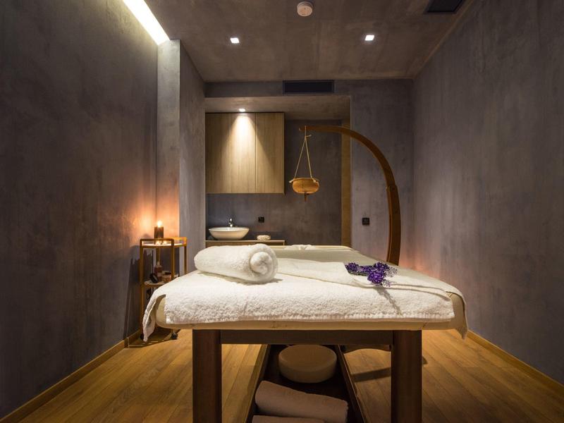 Schlichte Massageliege mit weißer Decke, Holzboden, graue Wände und gedämpftes Licht.