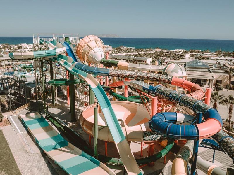 Wasserpark mit mehreren bunten Rutschen, Palmen und Blick auf das Meer im Hintergrund.