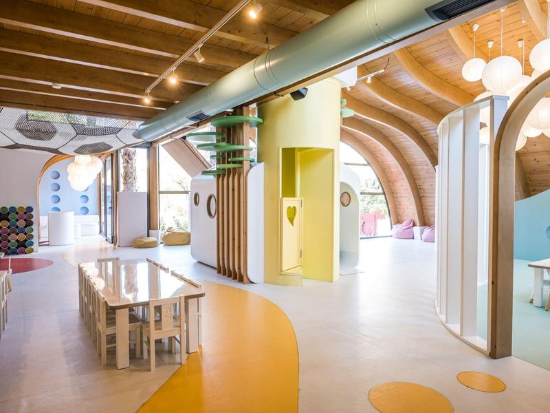 Heller, moderner Kindergartenraum mit bunten Böden, Holzmöbeln und geschwungenen Deckenformen.