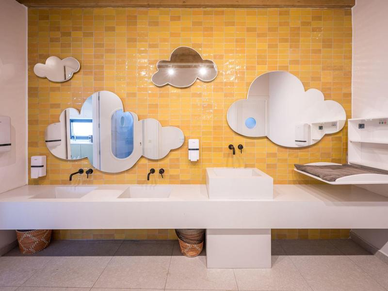 Moderner Waschbereich mit weißen Waschbecken, geschwungenen Spiegeln und gelber Wand.