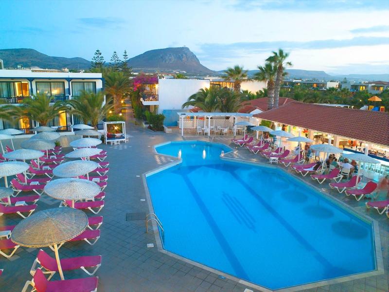 Großer blauer Pool mit rosa Liegestühlen und Sonnenschirmen, umgeben von Gebäuden und Bergen im Hintergrund.