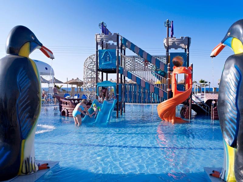 Wasser-Spielplatz mit Rutschen und großen bunten Pinguinfiguren am Rand eines Planschbeckens.