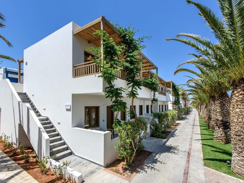 Weißes mehrstöckiges Gebäude mit Balkon, Palmen und Weg daneben unter blauem Himmel.