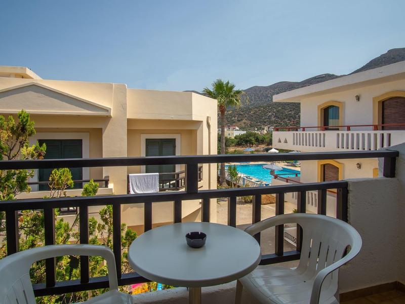 Balkón s stolem a židlemi s výhledem na bazén a sousední budovy pod jasnou oblohou.