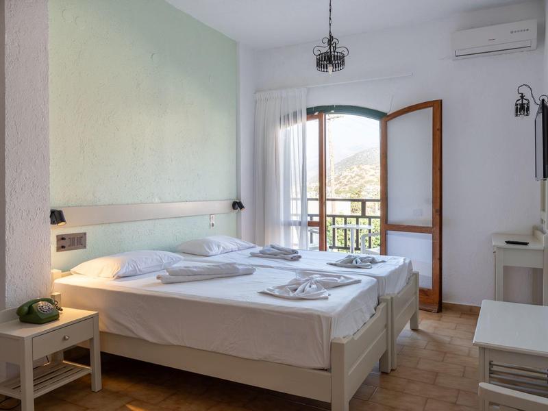 Světlý hotelový pokoj s dvěma postelemi, balkonem a výhledem na zelené kopce.