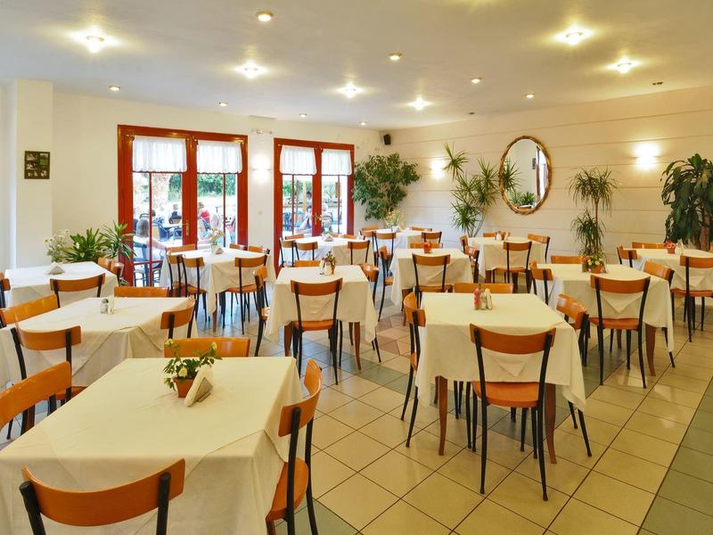 Světlá, prostorná jídelna s několika stoly a židlemi, rostlinami a velkými okny.