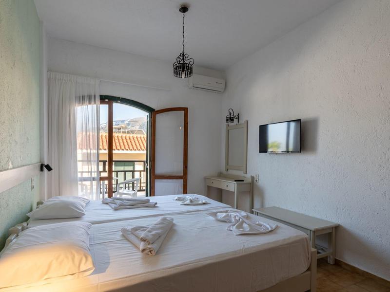 Světlá zařízená ložnice s manželskou postelí a balkonem s výhledem na hotel.