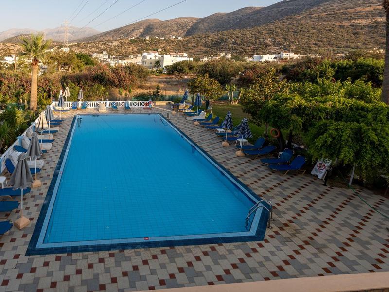 Obdélníkový bazén s modrými lehátky, obklopený stromy a horami v pozadí.