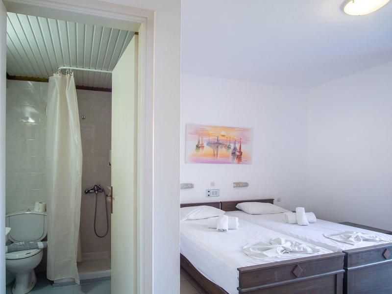 Světle hotelový pokoj s manželskou postelí, přilehlou koupelnou se sprchou a jednoduchým zařízením.