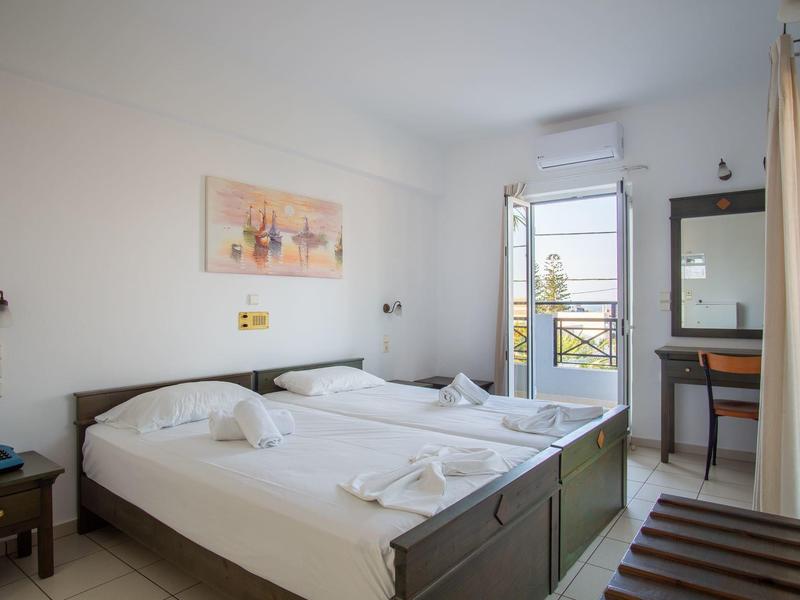 Světý hotelový pokoj se dvěma postelemi, nočním stolkem, psacím stolem a otevřeným oknem.