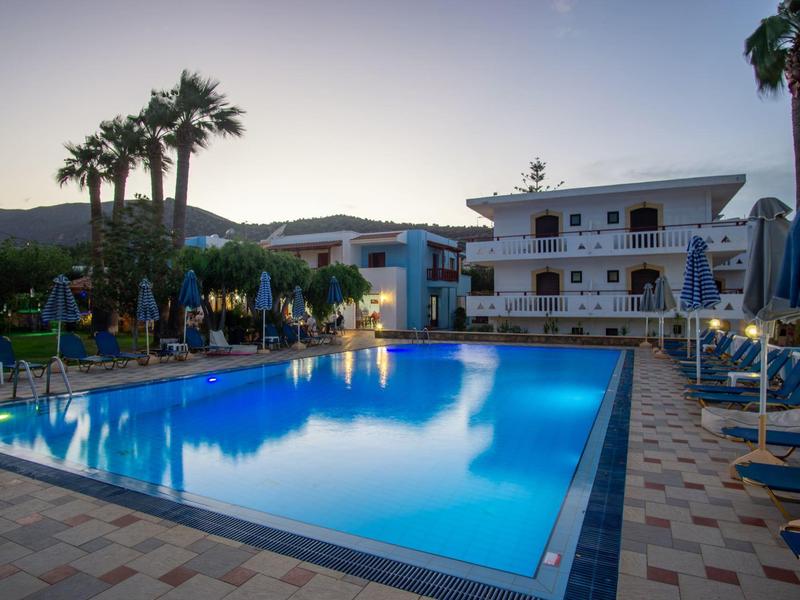 Pohled na osvětlený bazén s lehátky a palmami vedle hotelové budovy za soumraku.