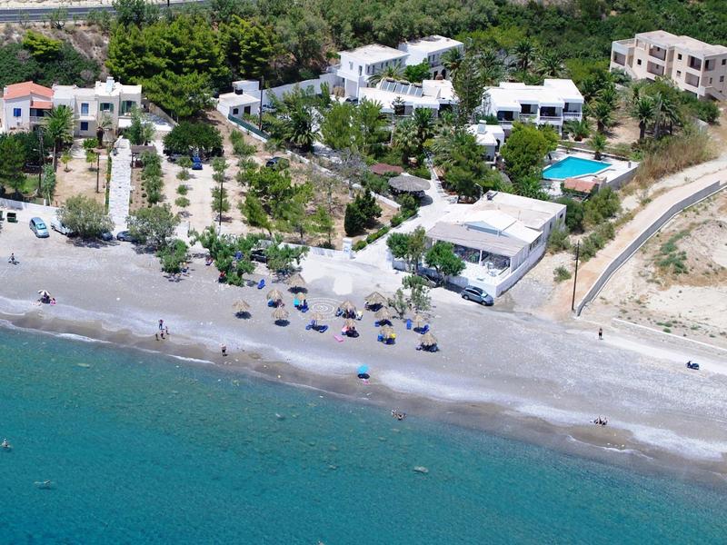 Vista aerea di una spiaggia con ombrelloni, ville bianche e acqua di mare limpida.