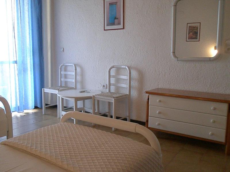 Camera d'hotel luminosa con letto matrimoniale, due sedie bianche, cassettiera e tende blu.