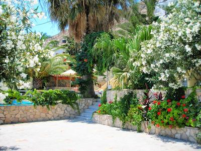 Zahrada s palmami, kvetoucími keři a kamennými zdmi pod modrou oblohou.