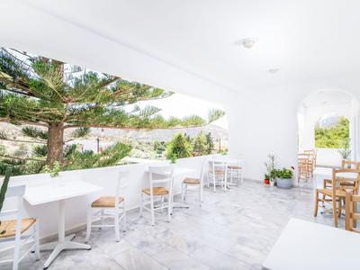 Světlá terasa s bílými zdmi, stoly a židlemi s výhledem na zelené stromy.