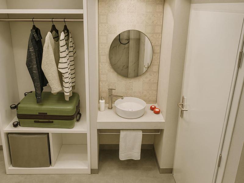Compacte hotelkamer met open kast, groene koffer en wastafel met ronde spiegel.