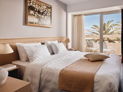 Jasny pokój hotelowy z podwójnym łóżkiem, białymi poduszkami i widokiem na taras z palmami.