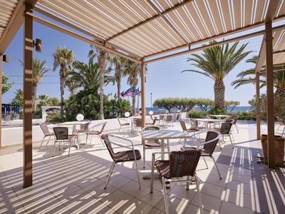 Terrace with tables and chairs under a pergola, palm trees and sea view in the background.