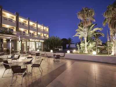 Area esterna dell'hotel con tavoli e sedie vuoti al crepuscolo, illuminata e con palme.