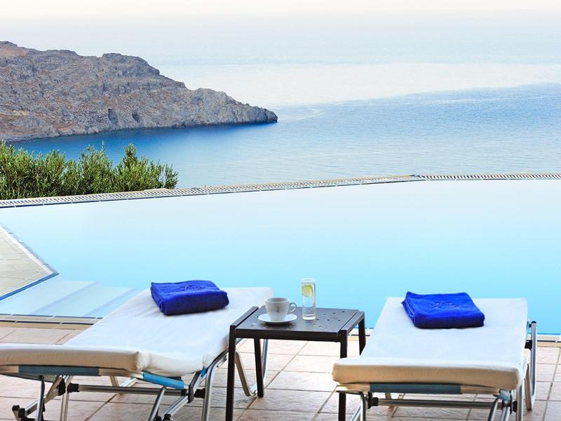 Piscina a sfioro con lettini e vista sul mare e sulle montagne sullo sfondo.