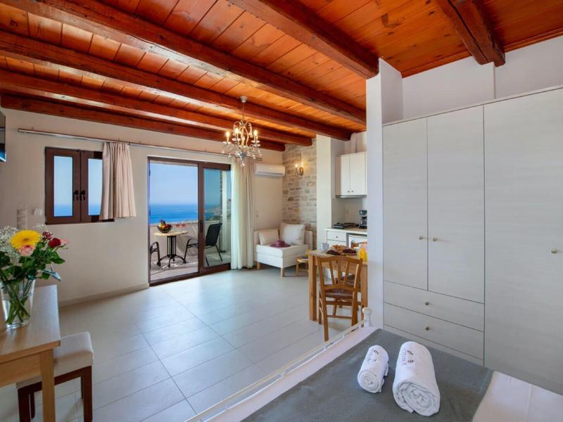 Camera d'hotel luminosa con soffitti alti con travi in legno, letto, tavolo, sedie e vista mare attraverso porte aperte.