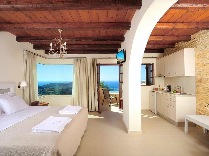 Camera d'hotel luminosa con soffitto a travi di legno, letto matrimoniale, angolo cottura e vista mare.