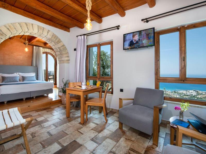 Camera d'albergo luminosa con travi in legno, poltrone, letto e grande finestra con vista mare.