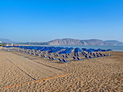 Leżaki z niebieskimi parasolami na pustej piaszczystej plaży z górami w tle pod jasnym niebem