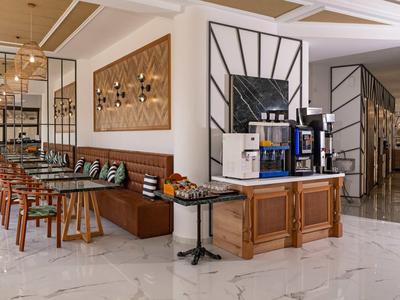 Nowoczesny hotelowy salon z drewnianymi krzesłami, marmurowymi płytkami i automatem do kawy samoobsługowym.