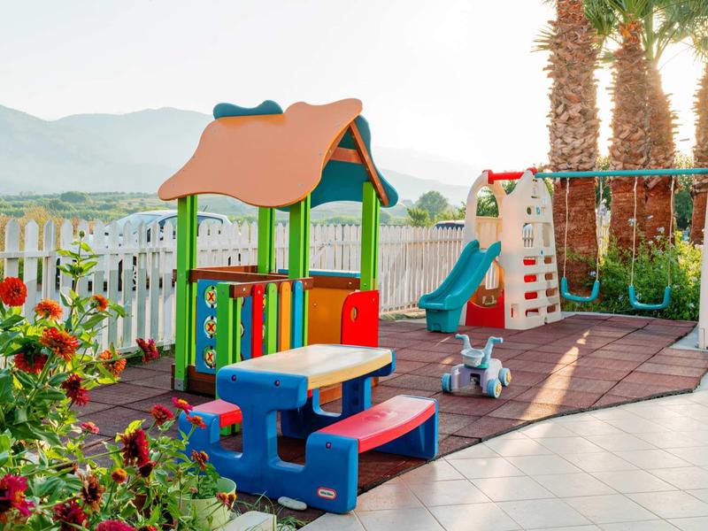 Bunter Spielplatz mit Rutsche, Kletterwand und Picknicktisch, umgeben von Palmen und Bergen.