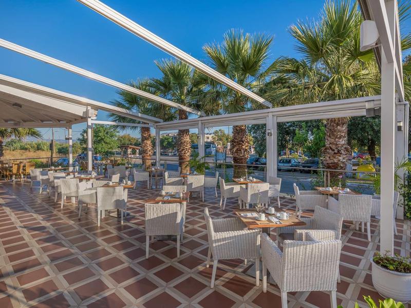 Freiluft-Terrasse mit weißen Stühlen, Holztischen und Palmen im Hintergrund bei Sonnenschein.