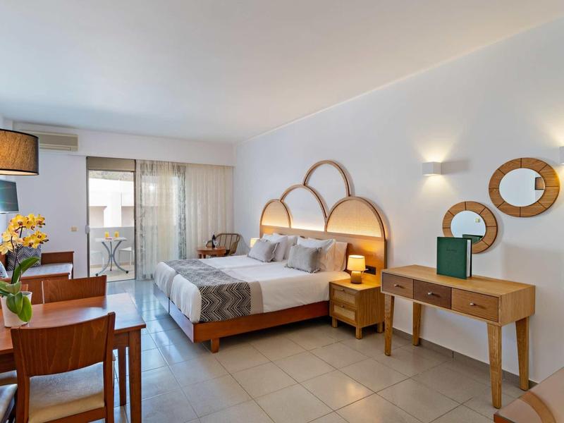 Helles Schlafzimmer mit Doppelbett, Holzmöbeln, zwei runden Spiegeln und großem Fenster mit Vorhang.