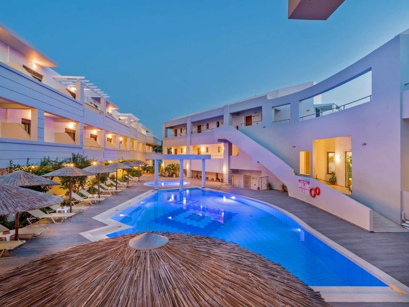 Moderner Hotelkomplex mit beleuchtetem blauen Pool, Sonnenliegen und weißen Gebäuden bei Dämmerung.